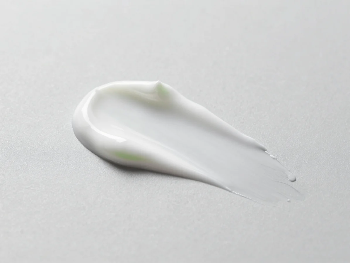 Tight texture study of a smooth white lotion swatch on light gray, soft diffused daylight, restrained highlights, editor